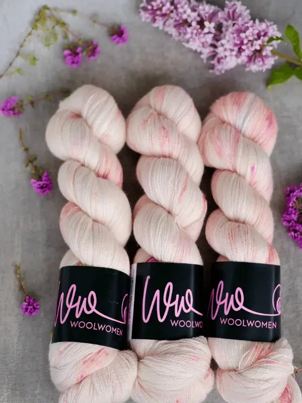 Three skeins of WoolWomen Merino Silk Lace in soft, light pink with speckles are arranged side by side on a gray surface, surrounded by small purple flowers and lilac blossoms.