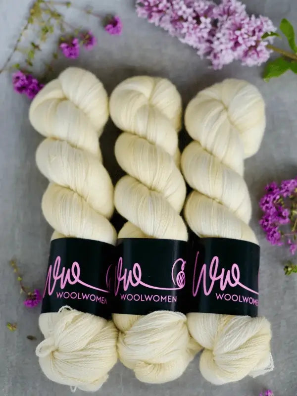 Three skeins of WoolWomen Merino Silk Lace in creamy white are arranged side by side on a gray surface, surrounded by sprigs of purple flowers and lilacs.