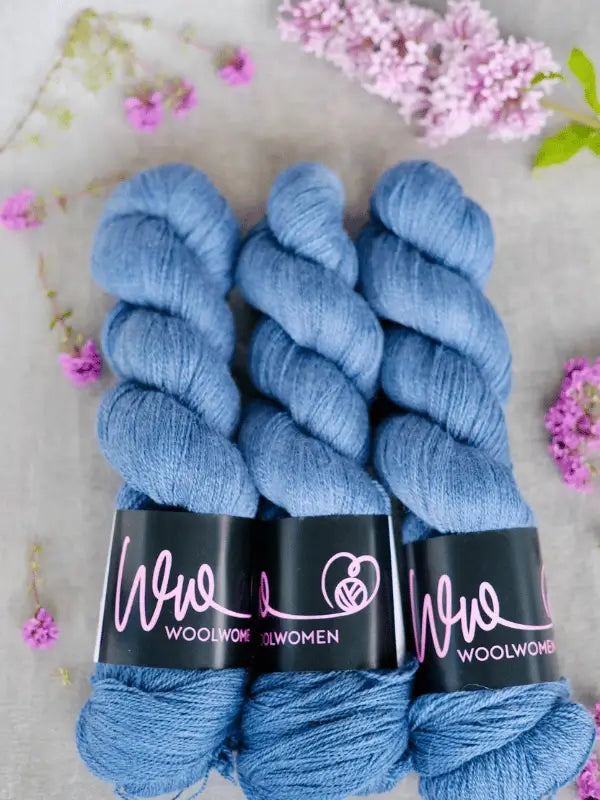 Three skeins of blue WoolWomen Merino Silk Lace yarn, crafted from käsinvärjätty merinovilla, are laid side by side on a light surface and surrounded by small purple and pink flowers.