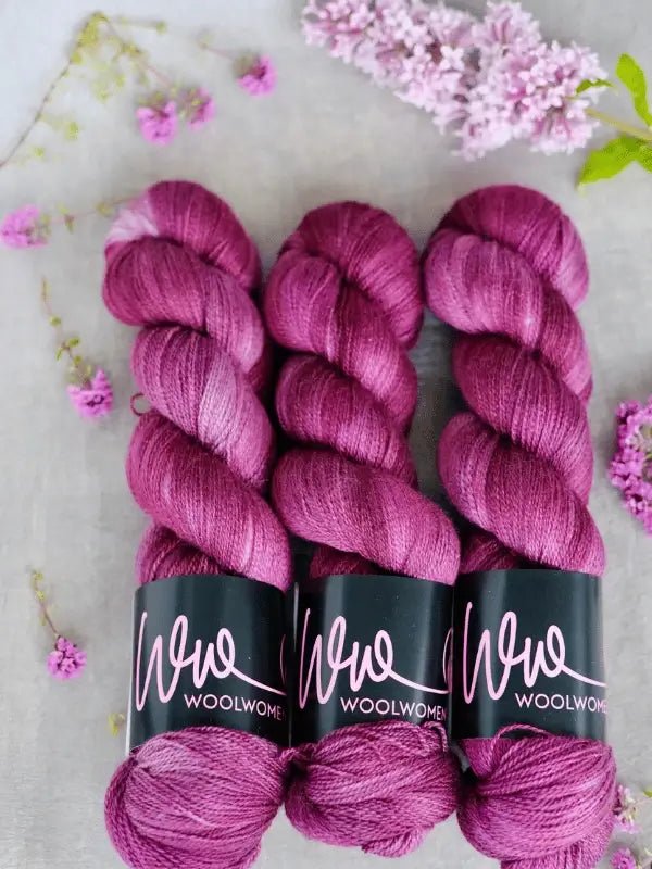 Three skeins of magenta WoolWomen Merino Silk Lace are arranged side by side on a light surface, surrounded by small pink and purple flowers with lilac blossoms in the background.