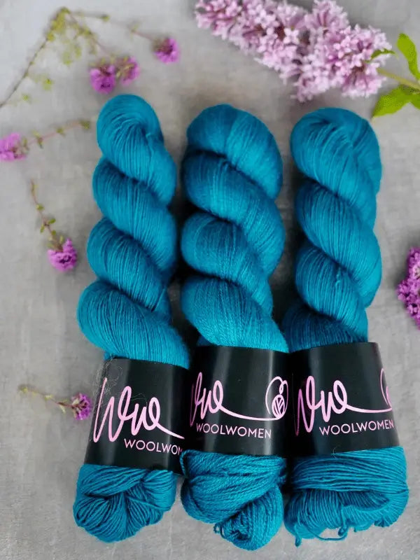 Three skeins of WoolWomen Merino Silk Lace yarn in teal blue are arranged side by side on a gray surface, with sprigs of purple flowers scattered around them.