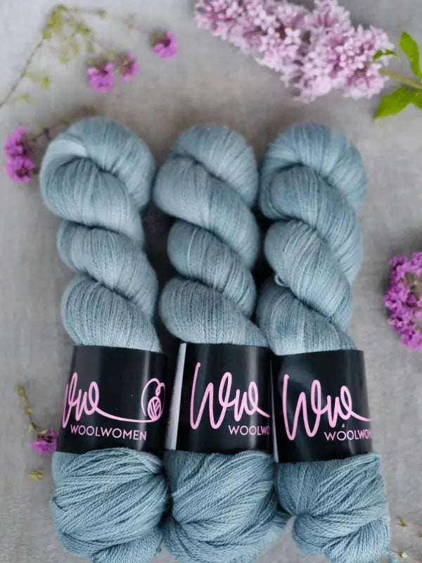 Three skeins of WoolWomen Merino Silk Lace yarn in light blue are side by side on a gray surface, surrounded by small pink and purple flowers.