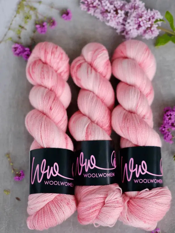 Three skeins of WoolWomen Merino Silk Lace in soft pink are neatly arranged side by side, with sprigs of purple flowers scattered around them on a light surface.