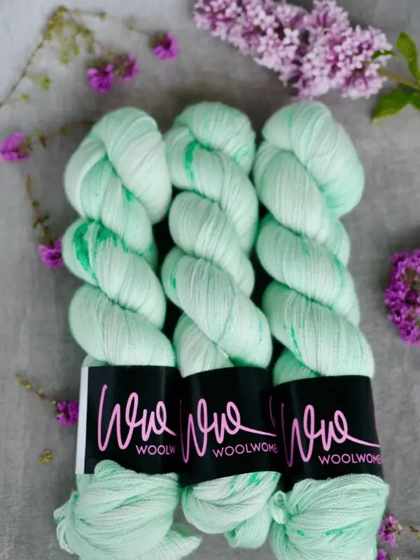 Three skeins of WoolWomen Merino Silk Lace yarn with subtle green specks, each labeled in pink on black wrappers, are arranged on a gray surface and surrounded by small purple flowers.