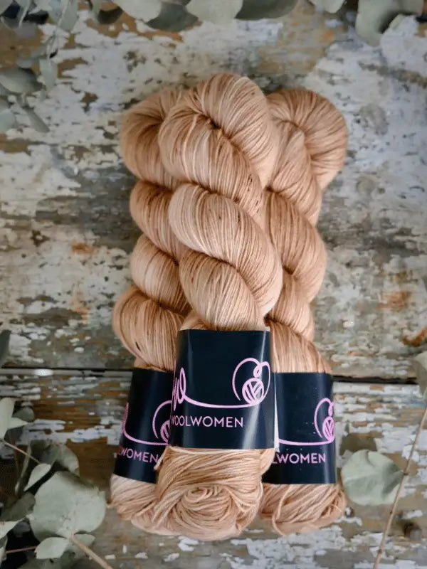 Two skeins of light beige WoolWomen Merino Sock yarn rest on a rustic wooden surface, surrounded by scattered dried leaves.