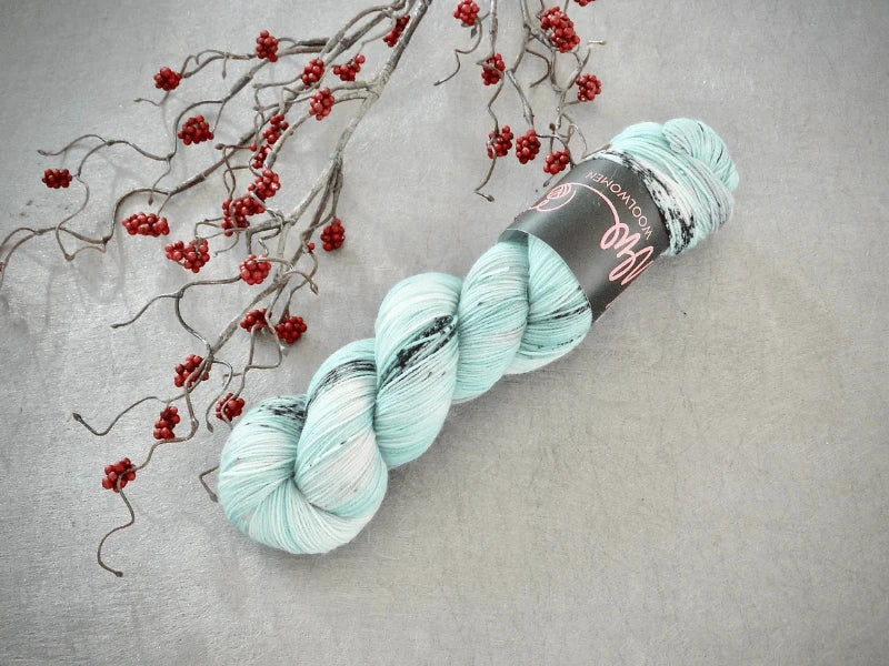 A skein of WoolWomen Merino Sock – Yhden Padan Ihme, light blue with gray and black specks, rests on a gray surface beside a branch with red berries.