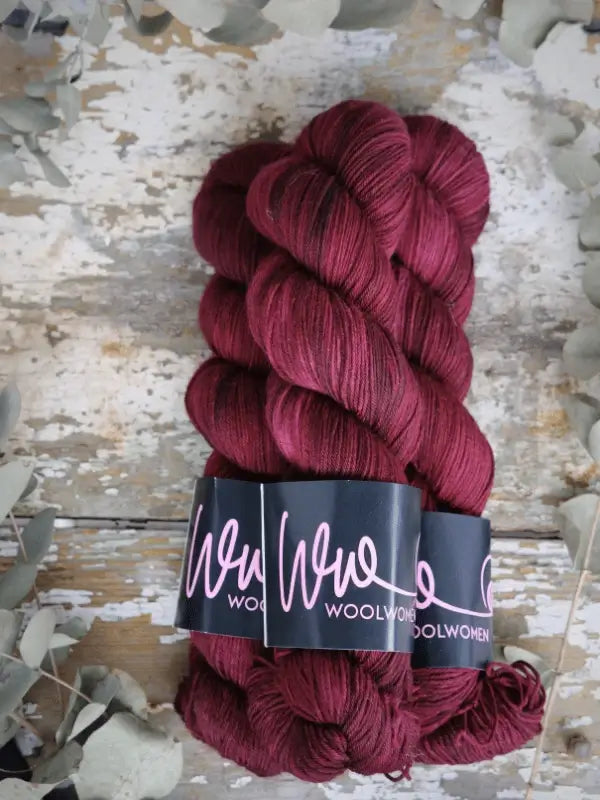 Three skeins of deep burgundy WoolWomen Merino Sock yarn are arranged on a rustic, weathered wooden surface, surrounded by pale dried foliage.