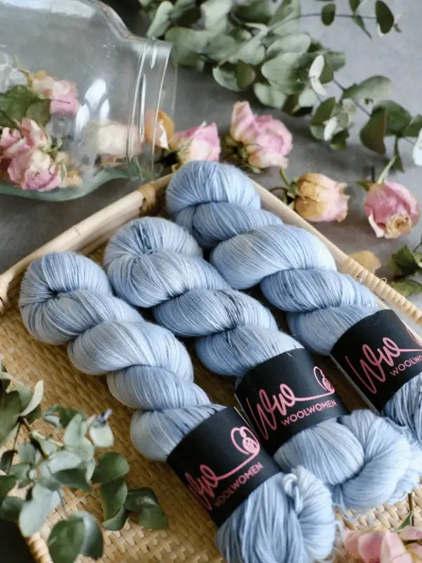 Three skeins of WoolWomen Merino Sock in pale blue with black labels rest in a woven tray, surrounded by dried rose petals and eucalyptus leaves scattered from a tipped glass jar.