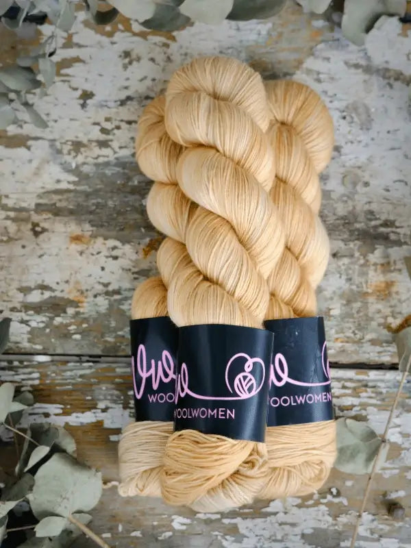 Two skeins of WoolWomen Merino Sock yarn in light beige rest on a rustic wooden surface, surrounded by dried leaves.