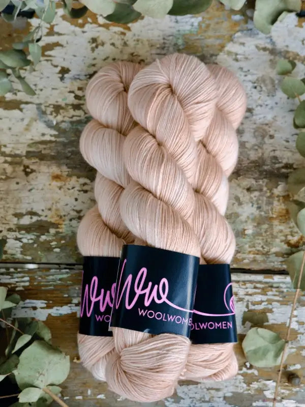 Two skeins of light beige WoolWomen Merino Sock yarn are side by side on a rustic wooden surface, surrounded by sprigs of greenery.