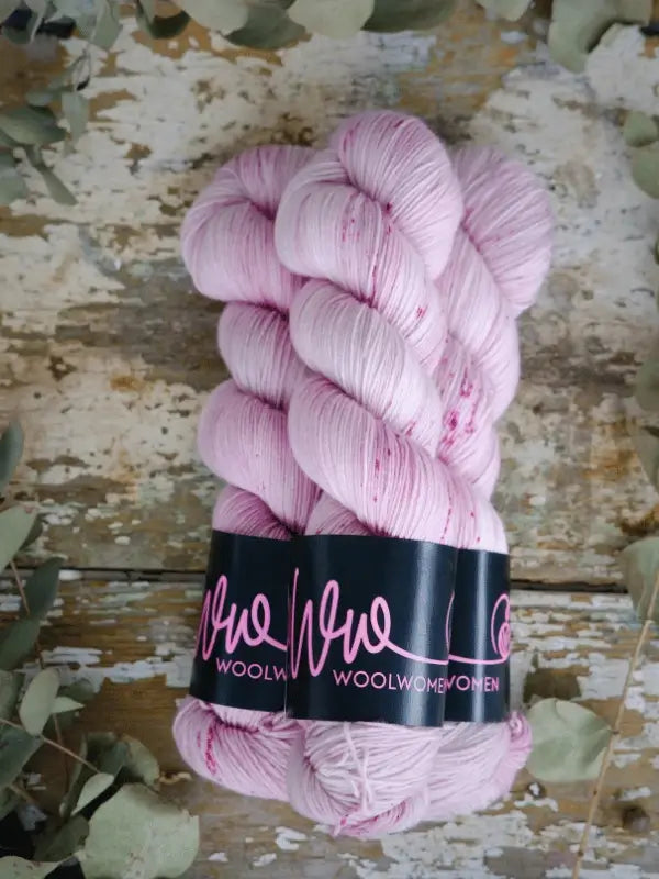 Three skeins of WoolWomen Merino Sock yarn in pale pink, wrapped with black labels, are arranged on a rustic wood surface with greenery. This käsinvärjätty lanka features subtle speckles for added charm.