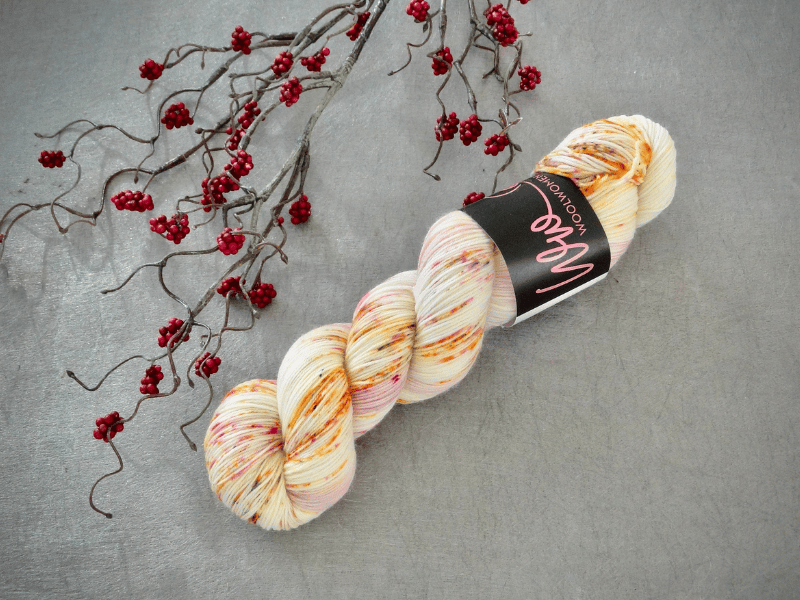 A skein of WoolWomen Merino Sock – Yhden Padan Ihme, käsinvärjätty lanka with orange, yellow, and pink speckles, rests on a gray surface beside a twig adorned with small red berries.