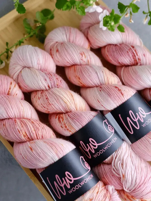 Three skeins of WoolWomen merino sock - Summer Collection yarn in pink and white speckles, each with a black Wool Women label, are neatly arranged on a wooden tray beside green leaves and small white flowers.