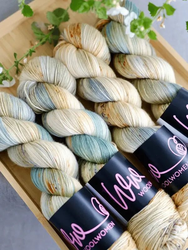 Four skeins of WoolWomen Merino Sock - Summer Collection yarn in cream, blue, green, and tan are arranged in a wooden tray with greenery at the corner. Each skein features a black WoolWomen label.