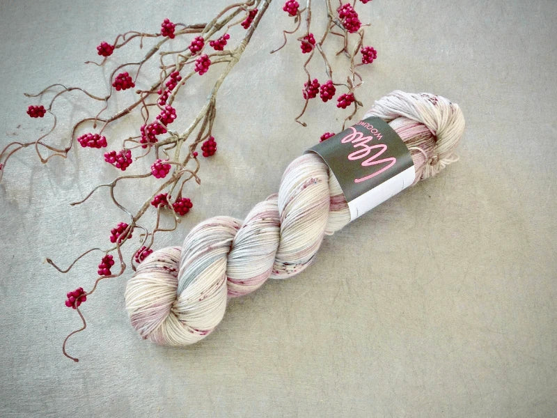 A skein of WoolWomen Merino Sock – Yhden Padan Ihme, a light-colored, speckled käsinvärjätty lanka with a label, rests on a gray surface next to a branch with small red berries.