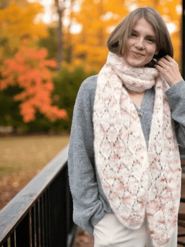 A person with light brown hair stands on a wooden deck, wearing a gray sweater and a chunky white and pink scarf knitted from the WoolWomen MinMax huiviohje pattern. Autumn trees with orange leaves appear in the background.
