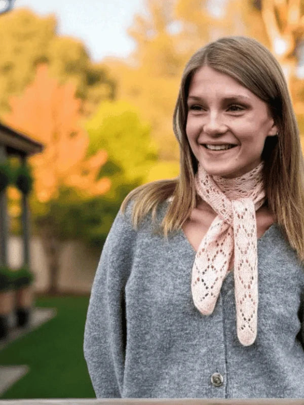 A smiling young woman with straight light brown hair wears a gray cardigan and a light pink lace WoolWomen MinMax huiviohje scarf, standing outdoors with trees and a building blurred in the background.