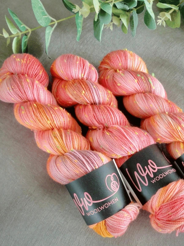 Three skeins of WoolWomen neon tweed—pink, orange, and yellow variegated with gold specks—rest on a gray surface. Each superwash merinovilla Donegal-lanka skein has a black WoolWomen label with a pink yarn logo; green leaves appear above.