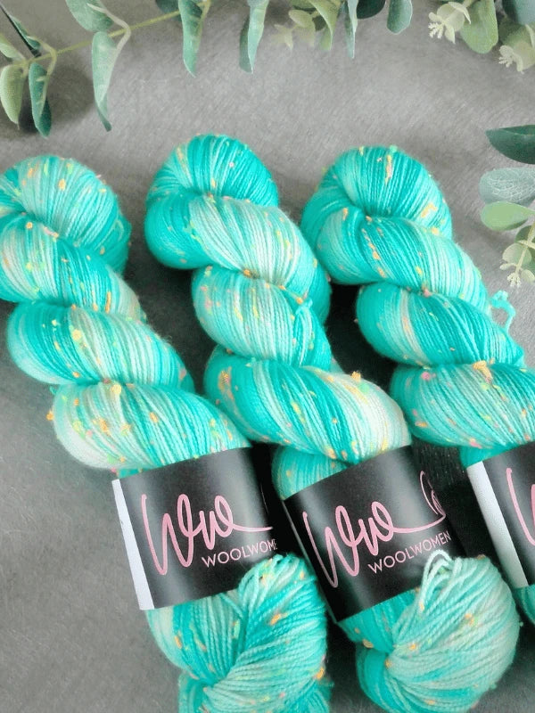 Three skeins of WoolWomen neon tweed yarn in turquoise, white, and yellow speckles are shown on a gray surface with green leaves. Each skein features a black Wool Women label.