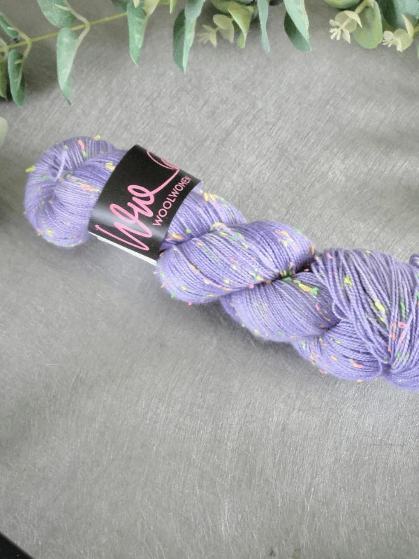A skein of WoolWomen neon tweed yarn in purple with multicolored specks, twisted and labeled, lies on a gray surface under green leafy branches. Made from superwash merinovilla Donegal-lanka for softness and durability.