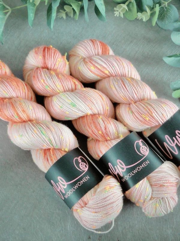 Three skeins of WoolWomen neon tweed yarn in pastel peach and pink with colorful neon flecks, arranged side by side on a light surface with greenery in the background.