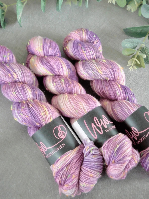 Three skeins of purple, lavender, and cream speckled WoolWomen neon tweed yarn lie side by side on a gray surface, each with labels reading WoolWomen. Green leaves are visible at the top of the image.