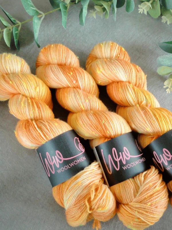 Three skeins of WoolWomen neon tweed yarn—a vibrant orange and yellow variegated superwash merinovilla with black labels—are arranged side by side on a gray surface, accented by green leafy branches in the background.