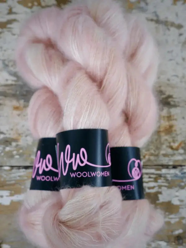 Two skeins of soft, light pink WoolWomen Silk Mohair yarn with black labels featuring the WoolWomen name in pink, displayed on a rustic wooden surface painted white and beige. This luxurious silk mohair yarn is perfect for any project that calls for an ylellinen neulelanka.