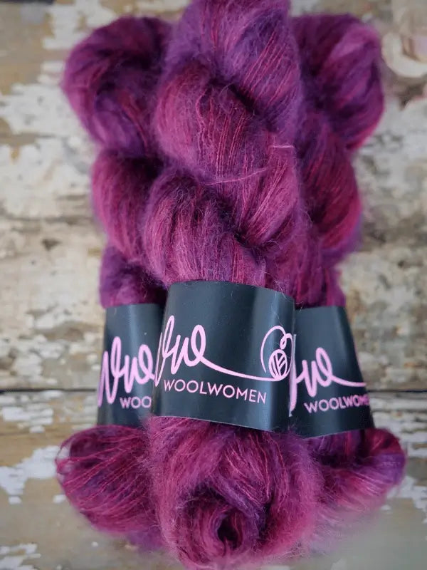 Three skeins of fluffy, deep magenta-purple yarn labeled WoolWomen Silk Mohair are stacked together on a distressed wooden surface. This ylellinen neulelanka, crafted from käsinvärjätty mohair, exudes both luxury and artistry in every strand.