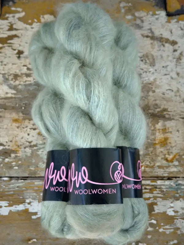 Three skeins of pale green, fluffy WoolWomen Silk Mohair yarn rest on a weathered, rustic wooden surface. This käsinvärjätty mohair and silkki-mohairlanka blend creates a truly luxurious neulelanka, ideal for sophisticated knitting projects.