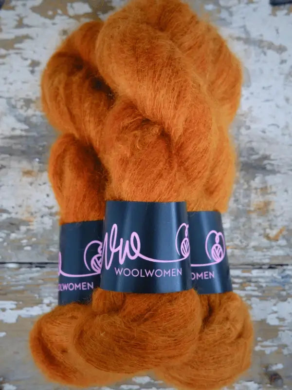 Two skeins of fluffy orange yarn with WoolWomen Silk Mohair labels are placed side by side on a distressed wooden surface. This ylellinen neulelanka features käsinvärjätty mohair, making it perfect for your most luxurious knitting projects.