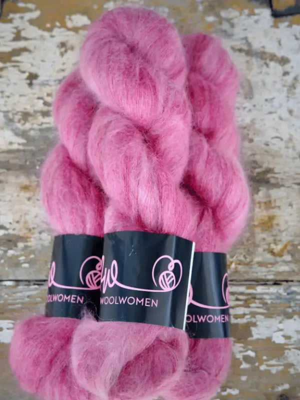 Three skeins of WoolWomen Silk Mohair—luxurious, fluffy pink yarn—are displayed on a rustic, weathered wooden surface. This silk mohair yarn blends the softness of hand-dyed mohair with premium quality, making it perfect for your next knitting project.