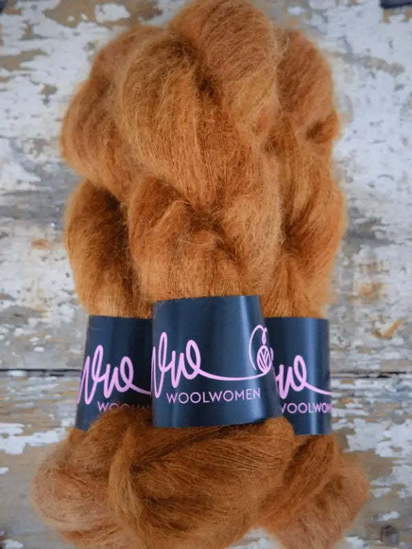 Three skeins of fluffy, rust-colored WoolWomen Silk Mohair are bundled with black bands labeled WOOLWOMEN in pink text, resting on a distressed wooden surface. This käsinvärjätty Silk Mohair lends an artisanal charm to your projects, making it perfect for those seeking unique and luxurious fibers.