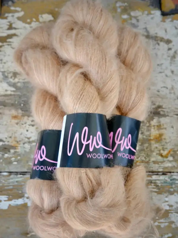 Three skeins of light tan WoolWomen Silk Mohair yarn, braided and labeled “WOOLWOOL” with black wraps and pink writing, rest on a rustic, weathered wooden surface. This luxurious silkki-mohairlanka is perfect for refined projects, bringing exceptional softness and elegance to your käsinvärjätty mohair creations.