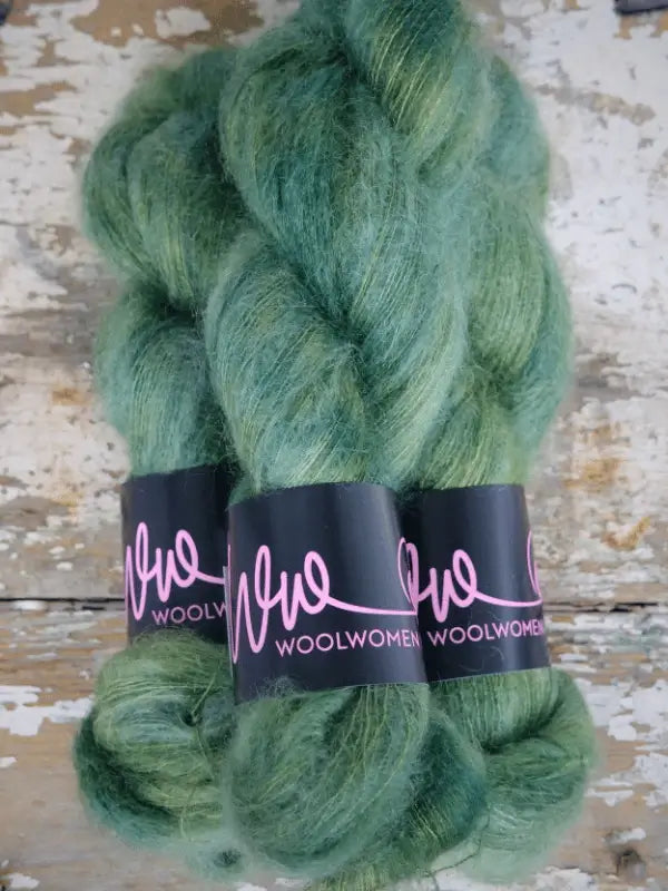 Three skeins of green WoolWomen Silk Mohair yarn, each with a black and pink WoolWomen label, displayed on a rustic, weathered wooden surface.