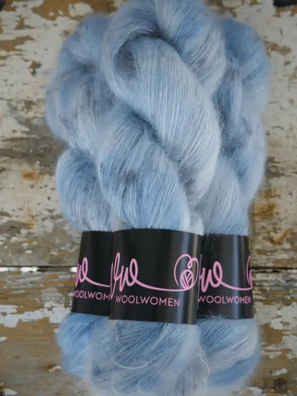 Three skeins of WoolWomen Silk Mohair in a soft, light blue shade, each with a fluffy texture and wrapped in a black label featuring "WoolWomen" in pink lettering, rest on a distressed wooden surface.
