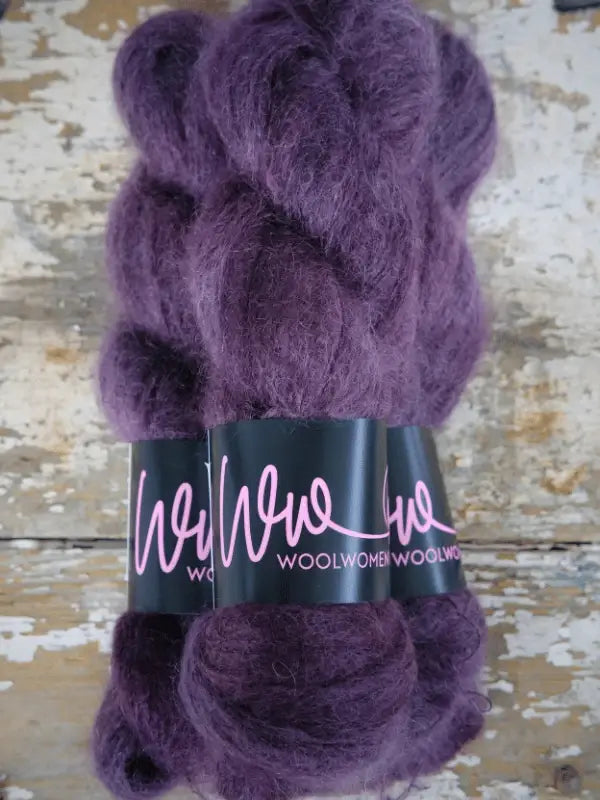Three skeins of fluffy purple yarn labeled WoolWomen Silk Mohair with black wrappers sit on a distressed wooden surface with white and beige peeling paint. This ylellinen neulelanka is a käsinvärjätty mohair blend, adding an elegant touch and luxurious softness to any knitting project.