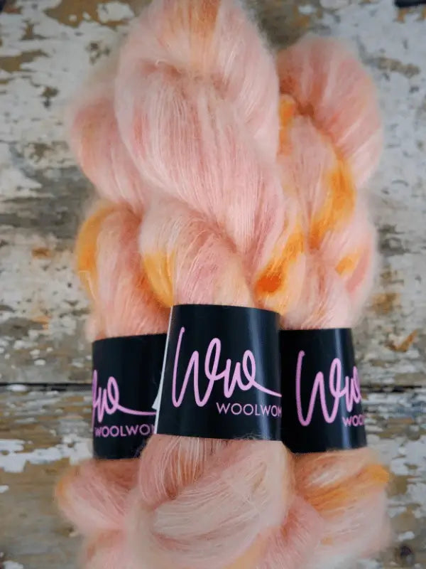 Three skeins of soft, fluffy yarn in pastel pink with orange accents, labeled “WoolWomen Silk Mohair,” are arranged side by side on a rustic, weathered wooden surface. This luxurious collection features käsinvärjätty mohair, making it the perfect ylellinen neulelanka for your next knitting project.