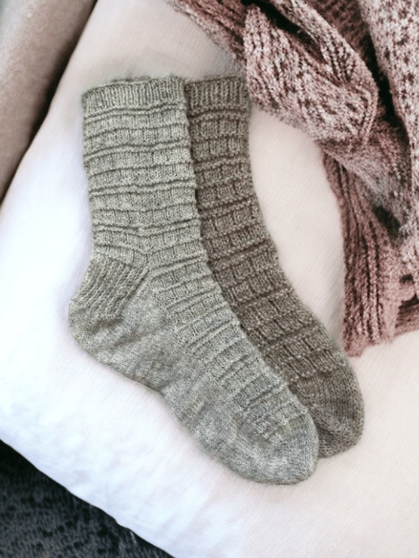 Two cozy, knitted socks in neutral tones—made with Aapo sukkaohje sport PDF and Lang Yarns Super Soxx 6 ply—lie side by side on a light blanket, with a pinkish textured throw partially visible nearby.