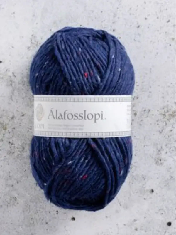 A skein of dark blue Istex Alafosslopi yarn, an iconic Icelandic wool with tiny flecks of color, wrapped in a white label, rests on a light gray textured surface.