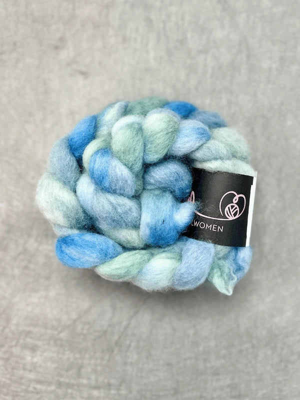A thick braid of soft, fluffy WoolWomen Suomalainen Kehruuvilla käsinvärjätty in blue and green shades is coiled into a bundle with a black label featuring a ball of yarn logo, set on a light gray textured surface.