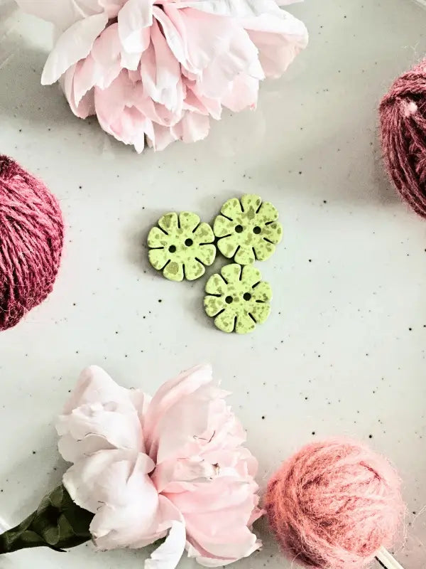 Three green Värikkäät kukkanapit 20mm (17700) are centered on a white background, framed by two pink peonies and two pink yarn balls in the corners, creating a balanced, yksinkertainen decorative display.