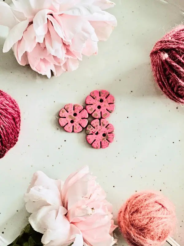 Three Värikkäät kukkanapit 20mm (17700) are arranged on a light surface, surrounded by pink yarn balls and two light pink peonies in opposite corners for a simple, charming effect.