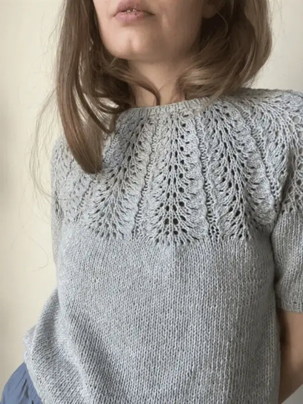 A woman wears a light gray short-sleeved knit sweater with a lacy yoke, made from Sandnes Garn Line and inspired by Le Knit Palme Tee neuleohje, ENG, standing against a plain background.