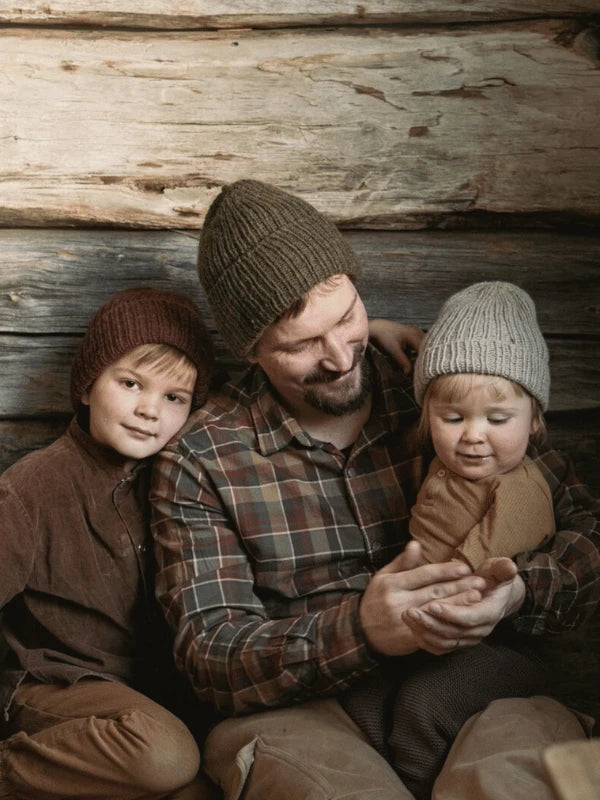 A man in a plaid shirt and knit hat sits with two kids, both in cozy knit hats, by a rustic wooden wall; their warm style suggests patterns from "Ronja Hakalehto, Kiertoteitä" as they smile and enjoy time together.