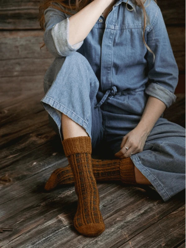 Wearing a blue denim jumpsuit and brown knitted socks, someone sits on a wooden floor, reflecting the cozy neuleohjeet style of Pohjois-Suomi—an ambiance captured in Ronja Hakalehto's "Kiertoteitä.
