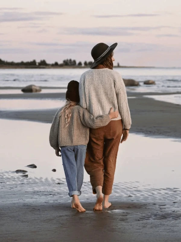 A woman and child in Ronja Hakalehto, Kiertoteitä sweaters and hats walk barefoot on a sandy North Finland beach, arm in arm, facing away as the sun rises or sets.