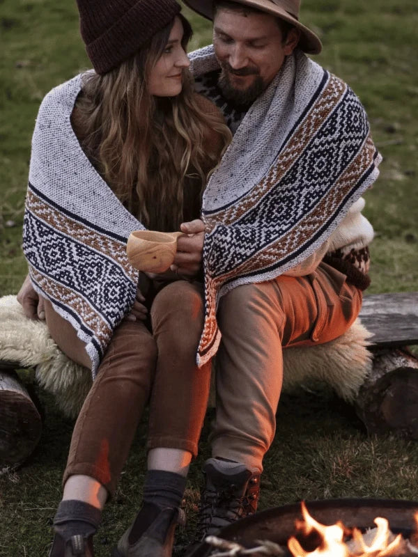 A couple sits by a campfire in Pohjois-Suomi, wrapped in the patterned Ronja Hakalehto, Kiertoteitä blanket and wearing cozy neuleohjeet-inspired clothes, gazing at each other on a log bench outdoors.