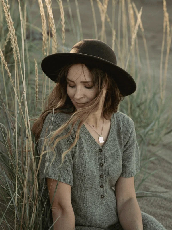 A woman in a grey dress and black hat sits in tall grass, softly illuminated by sunlight. Her gentle smile reflects the tranquil beauty of Pohjois-Suomi, capturing the serene mood of "Ronja Hakalehto, Kiertoteitä.