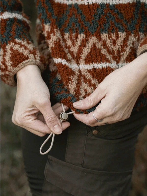 A person adjusts the drawstring at the hem of a patterned, textured sweater with geometric shapes from Ronja Hakalehto’s Kiertoteitä collection. Only their hands and torso are visible above dark pants.
